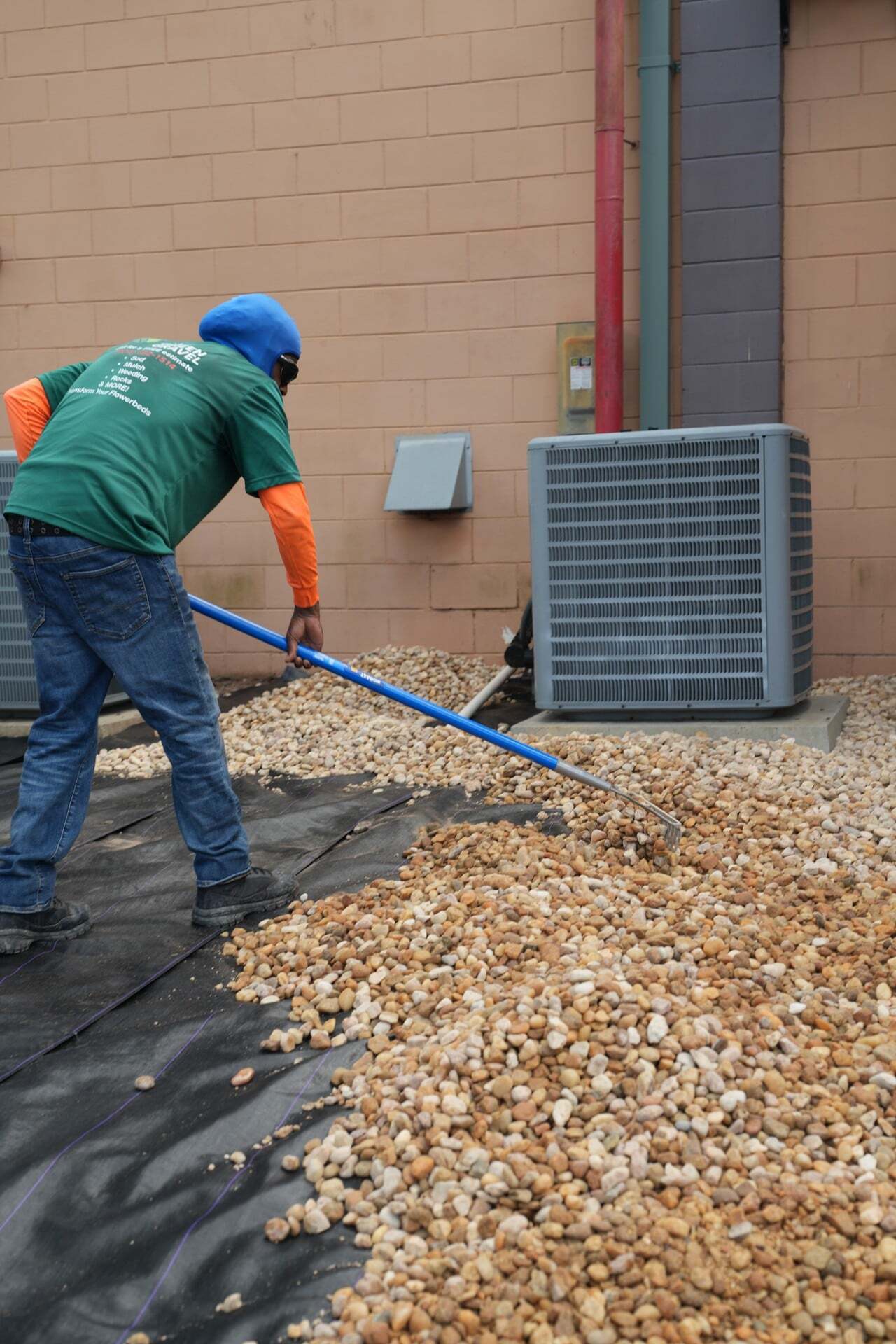 Crew member in a Green Gravel shirt raking a rock bed beside a commercial building